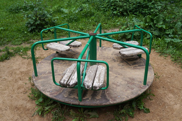 Small children's old carousel in a courtyard. Green knobs, shabby wooden seats, floor with peeling paint. Ground around, green grass in the background