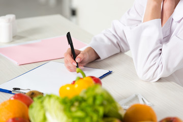 Obraz na płótnie Canvas partial view of dietitian in white coat writing in clipboard at workplace with fruits and vegetables on table