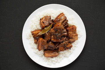 Homemade Filipino Adobo Pork with rice on a white plate over black background, top view. Flat lay, overhead, from above. Close-up.