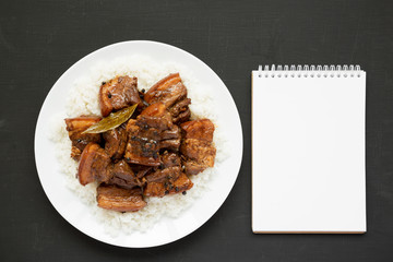 Homemade Filipino Adobo Pork with rice on a white plate, blank notepad over black surface, top view. Flat lay, overhead, from above. Copy space.