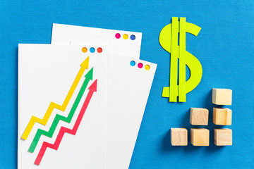 Inflation and financial concepts as profitability, taxes, stock market. Three paper arrows growing up and paper green dollar symbol with graph bar made with wooden blocks, on blue textured background.