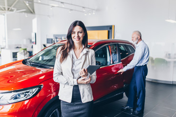 Saleswoman at the dealership showroom and man in the background choosing a new car for himself.