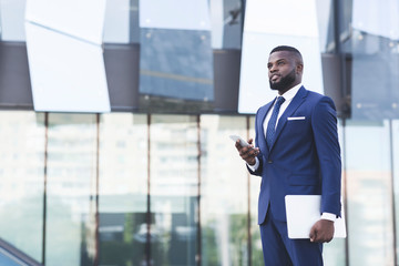 Fototapeta premium Afro Businessman Holding Phone And Laptop Standing In Urban Area
