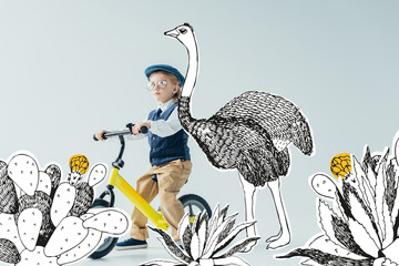 Thoughtful kid in retro vest and cap riding bicycle on grey background with fairy ostrich among...