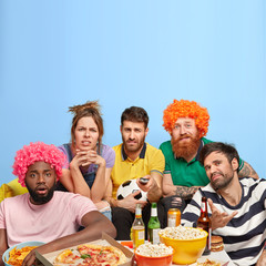Unhappy four men and one woman dissatisfied with final result of football match, upset favourite team lost game, surrounded with tasty pizza, popcorn, burgers and chips, gather together at home