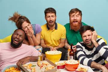Joyous group of multiethic millennial friends burst out of laughing, watch funny comedy on TV, have fun at home, eat fast food and drink beer, express positive emotions and feelings. Free time concept