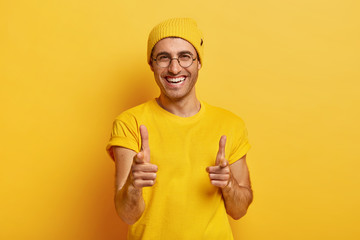 Portrait of happy man points fore fingers at camera, chooses you, looks gladfully at camera, smiles broadly, has white perfect teeth, friendly look, wears yellow clothes, poses indoor. Monochrome