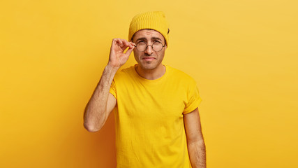 Discontent male model looks thoroughly through round glasses, has bad eyesight, tries to notice something little, touches frame of spectacles, has attentive look, dressed in casual yellow outfit