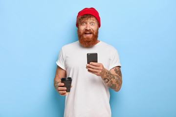 Positive millennial boy happy to get message from girlfriend, types answer, uses modern technologies during spare time, drinks takeaway coffee, wears red hat and white t shirt, has ginger beard