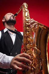 Fototapeta na wymiar Portrait of professional musician saxophonist man in suit plays jazz music on saxophone, red background in a photo studio, bottom view