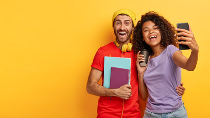 Cheerful joyful woman and man of different race make selfie photos on modern cellphone for blog, have happy expressions, going to university, dressed in casual clothes, take picture in front of camera