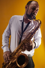 Fototapeta na wymiar Portrait of professional musician saxophonist man in white shirt plays jazz music on saxophone, yellow background in a photo studio, side view