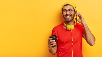 Indoor shot of cheerful millennial man speaks on mobile phone, looks away, holds takeout coffee makes call to friend dressed in stylish yellow hat red t shirt, enjoys spare time. People, communication