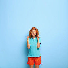 Fototapeta premium Hopeful ginger female model clenches fists with big desire to achieve goal, looks with intriguing expression, wears casual blue t shirt, red shorts, shows victory gesture, cannot wait for something
