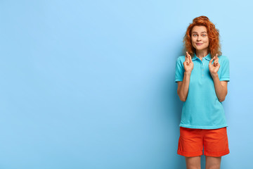 Praying beautiful woman crosses fingers for good luck, awaits faithfully for something important, hearing results, wears summer apparrel, isolated on blue background with empty space for promo