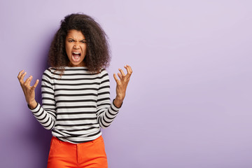 Dont bother me! Outraged Afro woman gestures angrily and screams at someone, has quarrel with boss, dressed in striped black and white jumper, stands agaist purple wall. Negative emotions concept