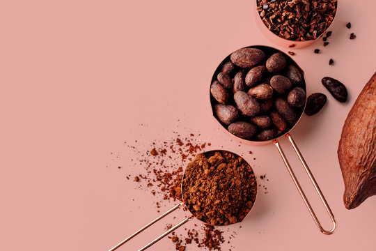 Rose gold measuring cups of cocoa beans, cacao nips, cocoa powder and cocoa pods on a pink background, flat lay healthy food concept