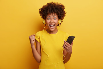 Positive hipster girl listens audio record with high volume, holds modern cell phone connected to headphones, clenches fist from good emotions, exclaims with joy, wears casual yellow t shirt