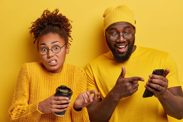 Glad surprised black guy points into screen of smartphone device, reads funny message content, puzzled curly haired woman holds takeout coffee, looks confusingly, dressed in yellow casual clothes
