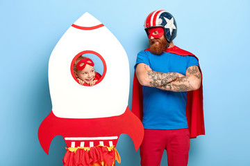 Horizontal shot of satisfied man wears headgear and red cape, keeps arms folded, looks at playful...