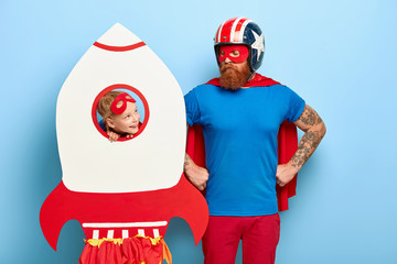 Serious father keeps hands on waist, wears headgear, blue shirt, red cloak looks at daughter play game together have fun at costume party with decoration. Lovely kid looks through rocket window at dad - Powered by Adobe
