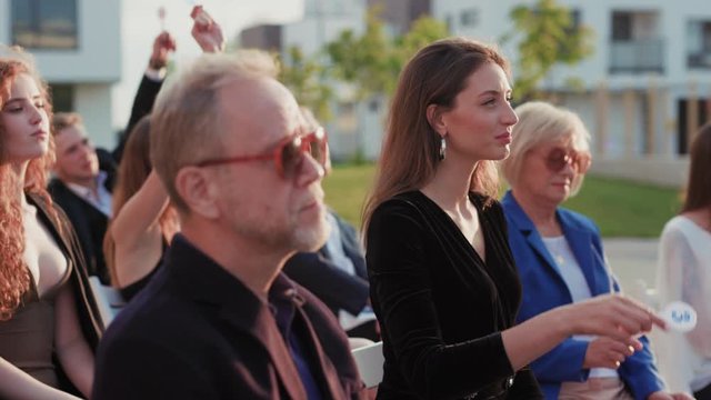 Side view attractive multi-age wealthy people bidding at modern auction. Ladies and gentlemen in formal wear sitting taking party in outdoor property sale event.