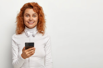 Fototapeta premium Attractive redhead girl has pleased look, holds phone, listens favourite music with headphones, has charming smile, wears white jumper with collar, poses indoor. People, technology, free time