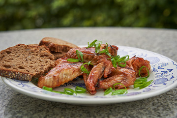 Hot chicken wings marinated in chili, soya sauce, ketchup and honey served on vintage rustic plate with whole grain bread and fresh chopped spring onion outside on the stone table in garden restaurant