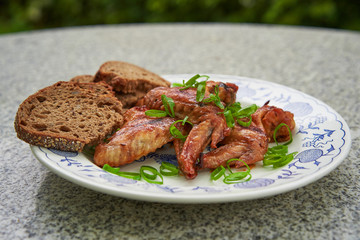 Hot chicken wings marinated in chili, soya sauce, ketchup and honey served on vintage rustic plate with whole grain bread and fresh chopped spring onion outside on the stone table in garden restaurant