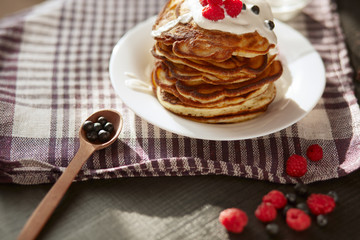 Image of cheese pancakes with sour cream and fresh berries, blackberry jam in wooden spoon, whipped cream on muffins top, served in white plate on with kitchen checkered dish towel, tasty dessert.