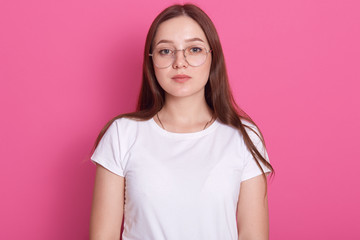 Fototapeta premium Close up portrait of beautiful young woman wearing spectacles and white casual outfit, feels happy, looks at camera, model posing isolated over pink studio wall, has long hair. People concept.