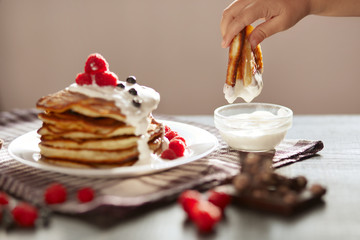 Side view of traditional family pancakes with sour cream and fresh berries isolated on dark background, woman's hand dipping muffin into sour cream, delicious dessert on checkered cotton towel.