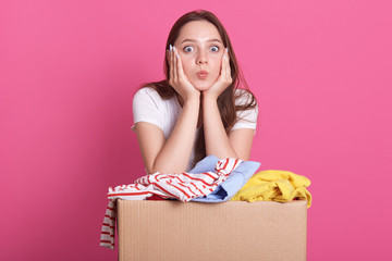 Studio shot of cheerful volunteer standing isolated over pink background in studio, near box with donated clothes, young attractive woman makes air kiss, looks directly at camera. Charity concept.