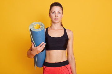 Sporty girl holding yoga mat in hands, wearing pants and black bra, looking directly at camera,...