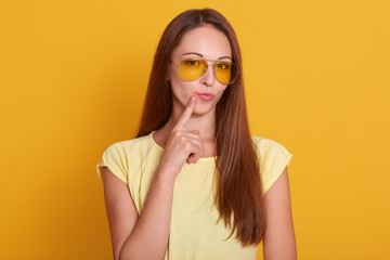 Close up portrait of insidious female keeps fore finger near lips, thinks about coming holidays, isolated on yellow background with blank copy space for advertisement or text, female with long hair.