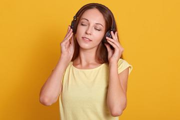 Close up portrait of beautiful Caucasian woman listening to music via headphone, relaxing while having free time, posing with closed eyes, dressed casual outfit, isolated over yellow studio background