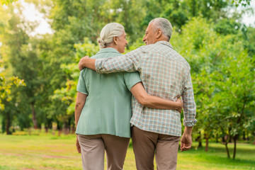 Rear view of elderly man and woman walking through a park with their arms around