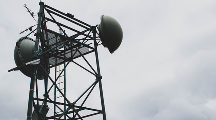 A cellular tower with satelite dishes, antennas and an access ladder against the cloudy white sky background with plenty of copy space.