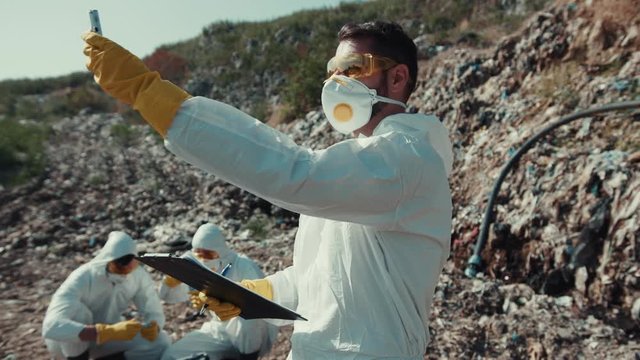 Recycling workers performing scientific research on landfill. Close-up young ecologist in suit estimating air pollution on huge waste dump.