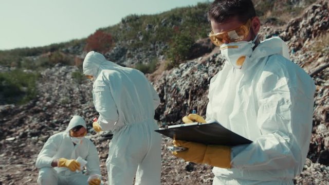 Ecology team working on landfill site collecting toxic garbage and estimating pollution. Close-up caucasian ecologist writing down notes standing near garbage rocks. Pollution concept.