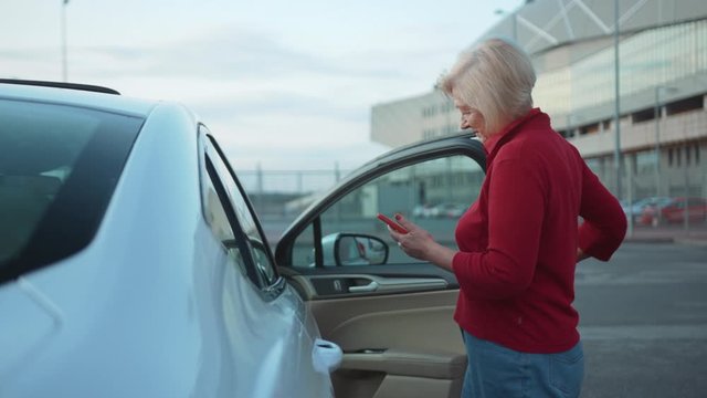 Happy senior woman stand near car use red cell phone gets in the car smile businesswoman blonde people smartphone cellphone communication internet search lady mobile browse network online slow motion