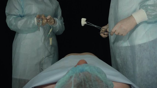Surgeons stand before operation or procedure. Nurse preparing drug injection with dropper. Patient lies on a surgical table in hospital. Doctor with scalpel and forcep is ready to start cut a patient
