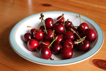 A bowl full of ripe organic red cherries with stalks on.  Red cherries sitting in a white bowl with a blue rim on a pine table.