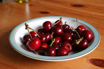 A bowl full of ripe organic red cherries with stalks on.  Red cherries sitting in a white bowl with a blue rim on a pine table.
