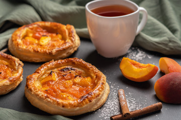 Homemade round puffs with peaches on a dark background with a linen napkin. Next to white cup with black tea, peach slices, cinnamon sticks, powdered sugar. Close up, horizontal orientation