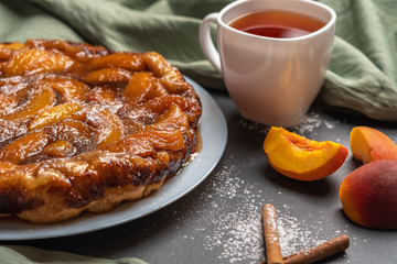 arte Tatin with peaches, caramel and powdered sugar close-up on a white plate on a dark background with a linen napkin. Next to the white Cup with black tea, peach slices and cinnamon sticks.