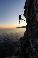 Silhouette of a Unrecognizable man rappelling down a steep cliff on the rocly ocean coast during a sunny summer sunset. Taken in Lighthouse Park, West Vancouver, British Columbia, Canada.