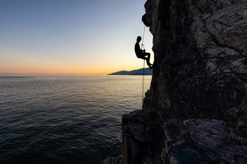 Silhouette of a Unrecognizable man rappelling down a steep cliff on the rocly ocean coast during a sunny summer sunset. Taken in Lighthouse Park, West Vancouver, British Columbia, Canada.