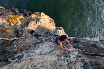 Adventurous Girl is Rock Climbing up a Steep Cliff during a summer sunset. Taken in Lighthouse Park, West Vancouver, British Columbia, Canada.