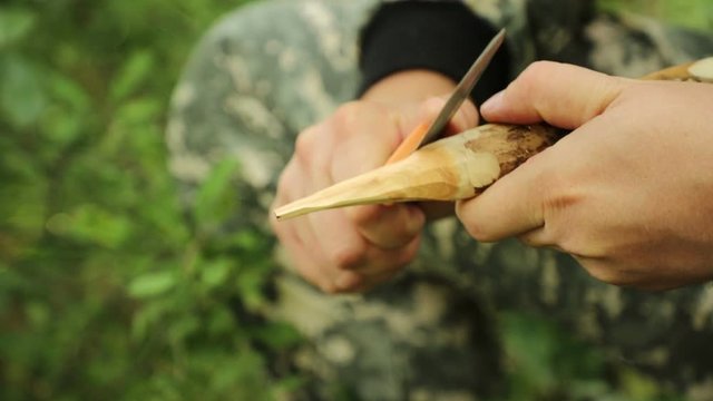 young handsome caucasian traveler in camouflage clothes, a cap and with a backpack, resting on a log  in a green forest and knife stabbing a stake from a stick, medium plan